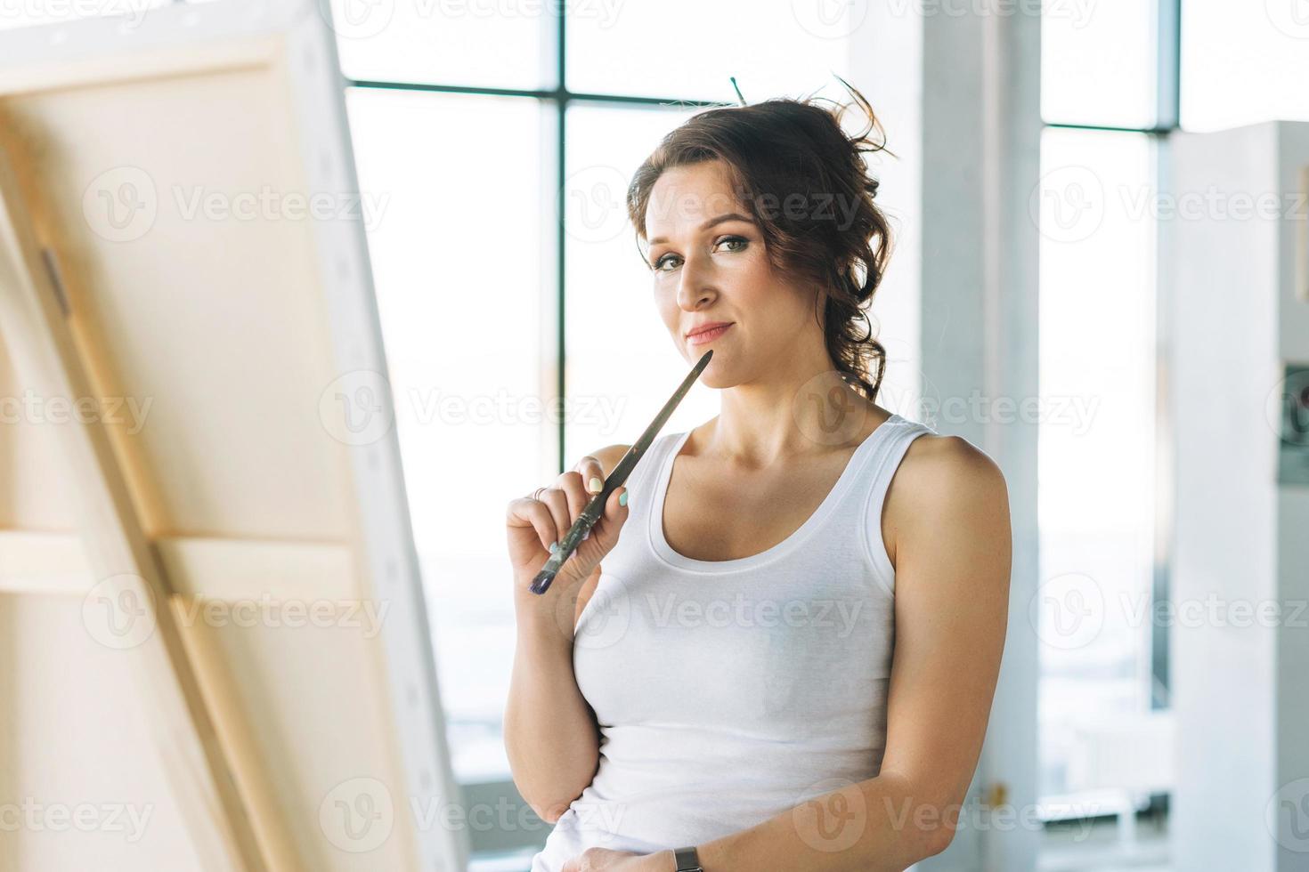 Pensando en una encantadora joven artista vestida de forma informal con pinceles frente al caballete en la galería de arte moderno foto