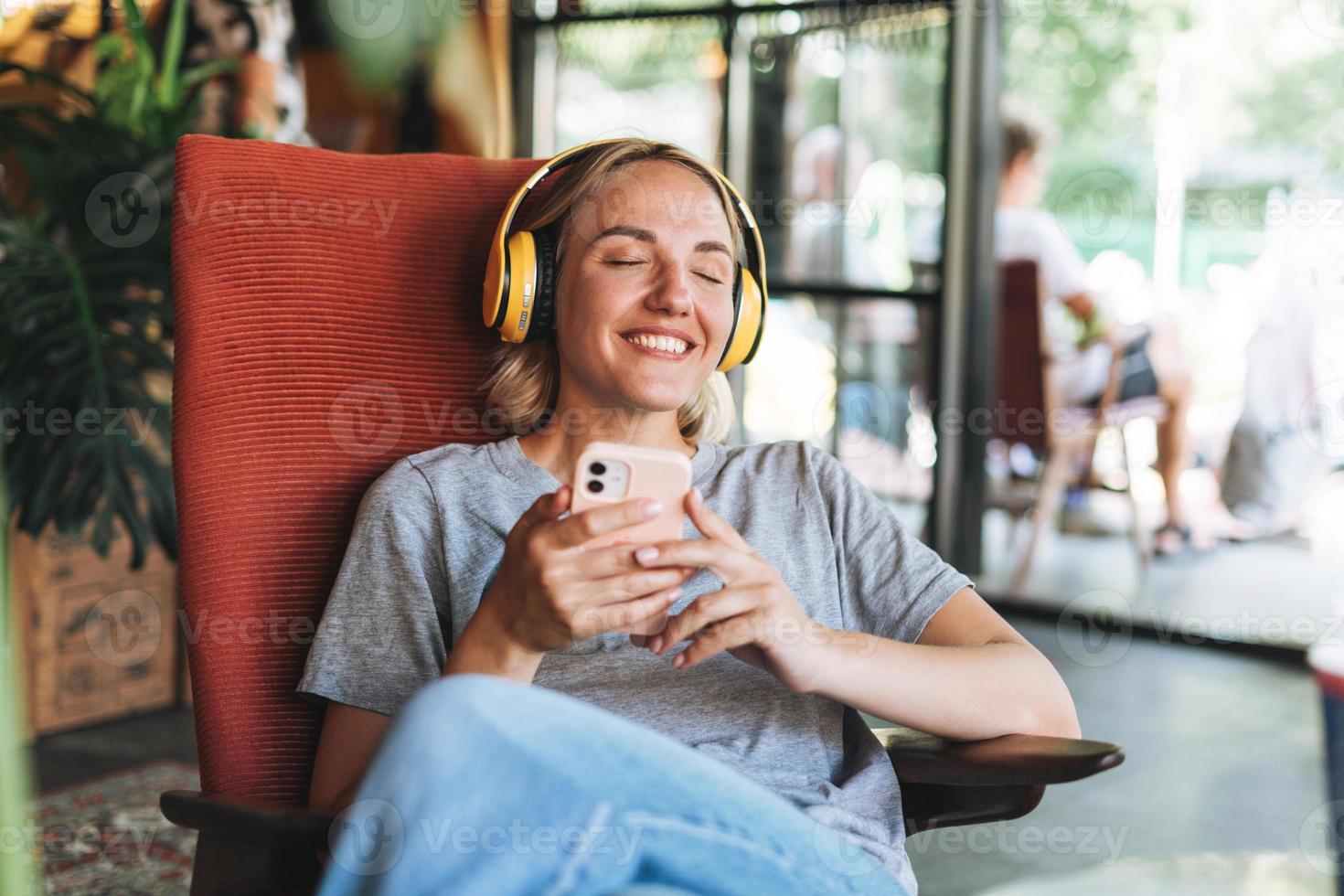 Young smiling blonde woman with close eyes in yellow headphones enjoys music with mobile phone sitting on chair at cafe photo