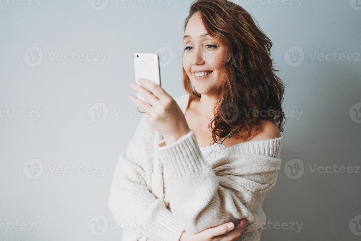 Attractive friendly smiling brunette woman middle age in cozy knitted cardigan using mobile isolated on grey background photo