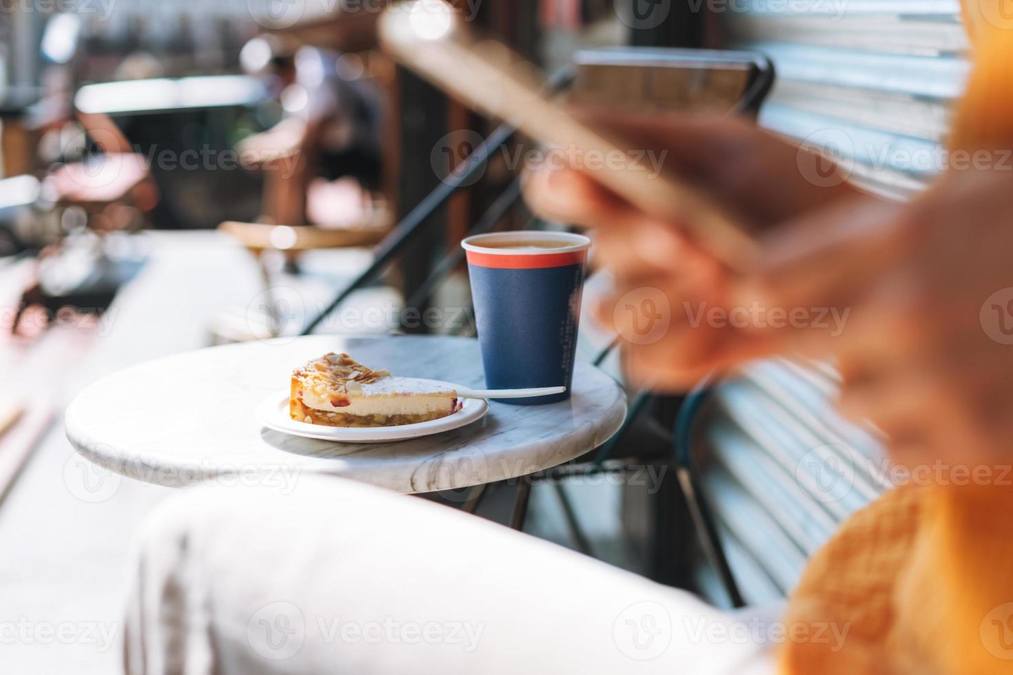 Paper cup with coffee and piece of cheesecake on table in outdoor cafe on background of blurred young woman with the mobile phon photo