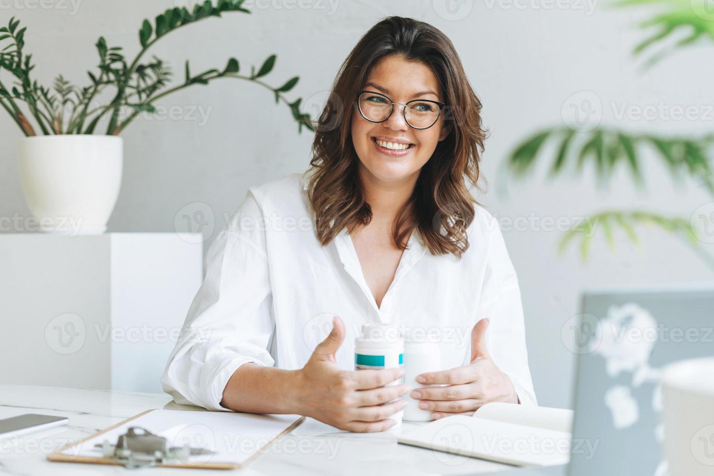 joven nutricionista morena sonriente de talla grande con camisa blanca que trabaja en una laptop en la mesa con una planta en la luminosa oficina moderna. el médico se comunica con el paciente en línea foto
