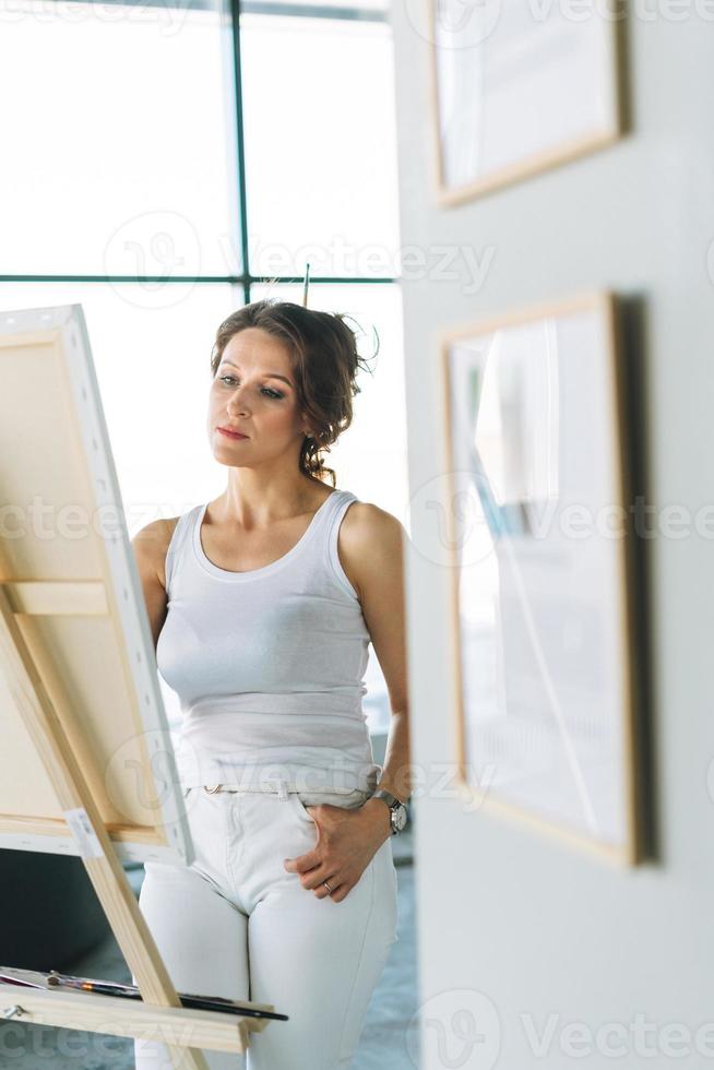 Thinking charming young woman artist in casual clothes with paint brushes in front of easel in modern art gallery photo