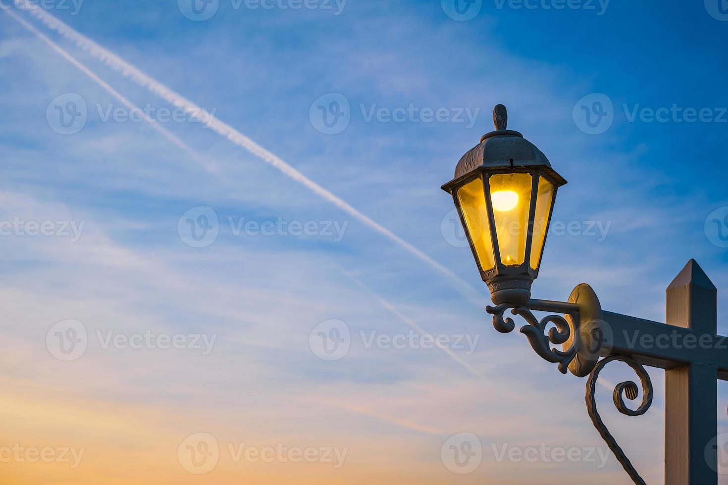 Electric lamp in retro style on a wooden pole. The lamp glows against the background of the sky with clouds during sunset. Idea for holiday background or postcard, copy space for text photo