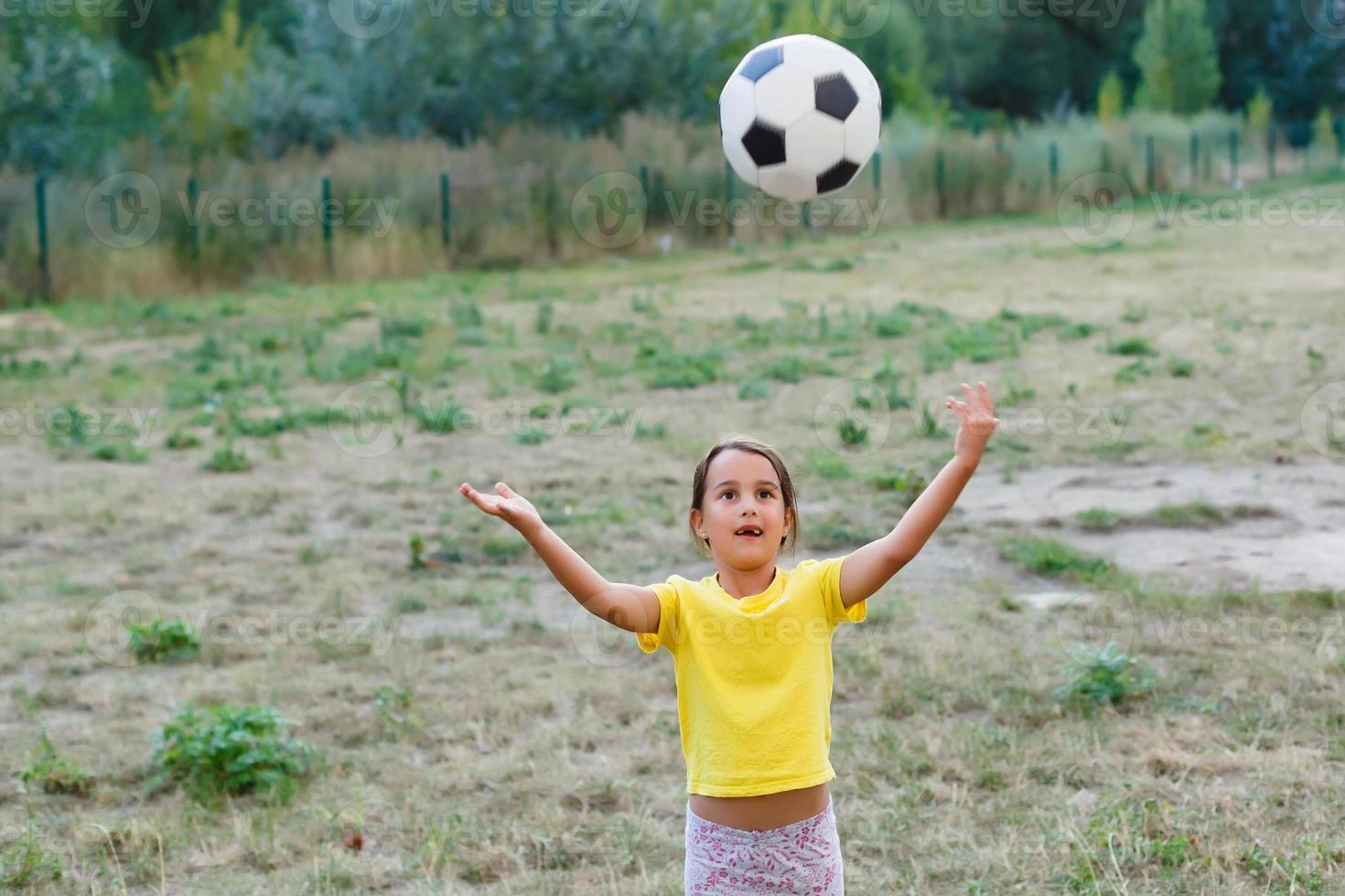 foto al aire libre de una linda niñita apoyada en una pelota de fútbol en hierba verde