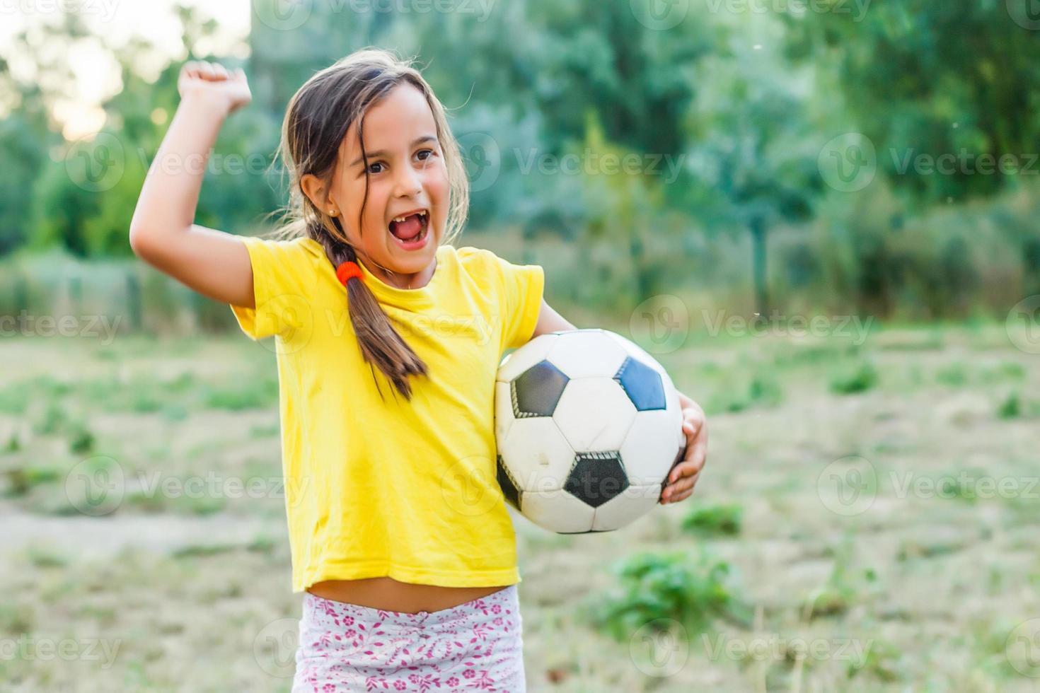 Outdoor photo of cute little girl leaning on soccer ball in green grass