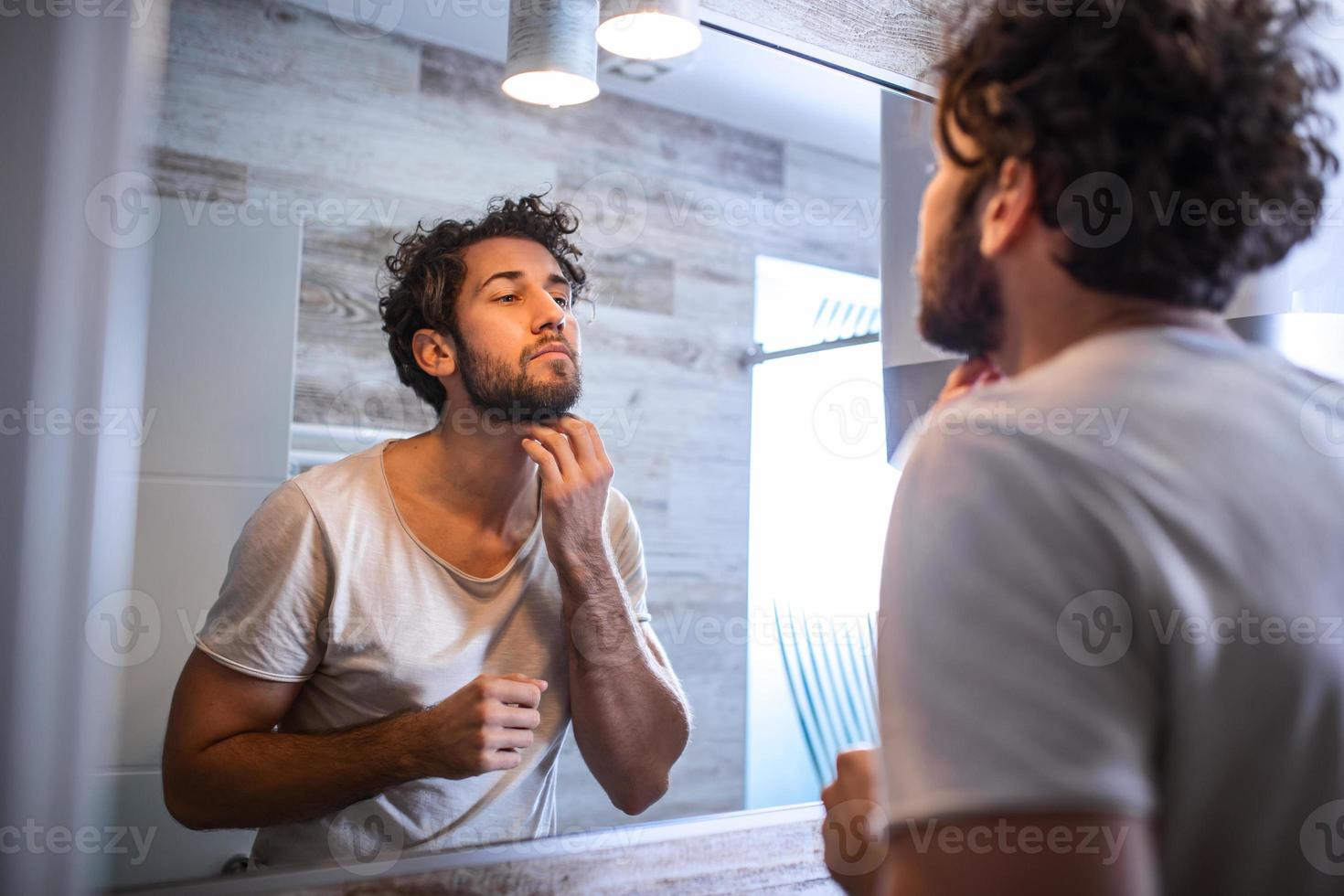 Reflection of handsome man with beard looking at mirror and touching face in bathroom grooming photo