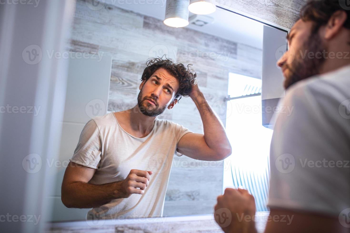 Handsome young man touching his hair with hand and grooming in bathroom at home. White metrosexual man worried for hair loss and looking at mirror his receding hairline. photo