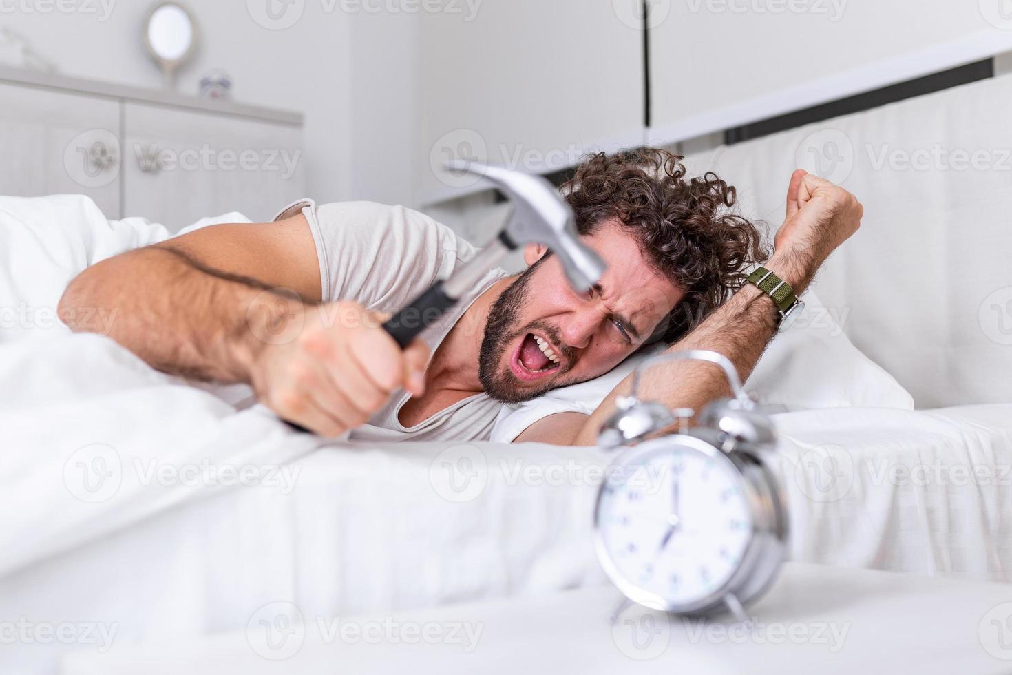 el joven intenta romper el despertador con un martillo, destruir el reloj. hombre acostado en la cama apagando un despertador con un martillo por la mañana a las 7 am. foto