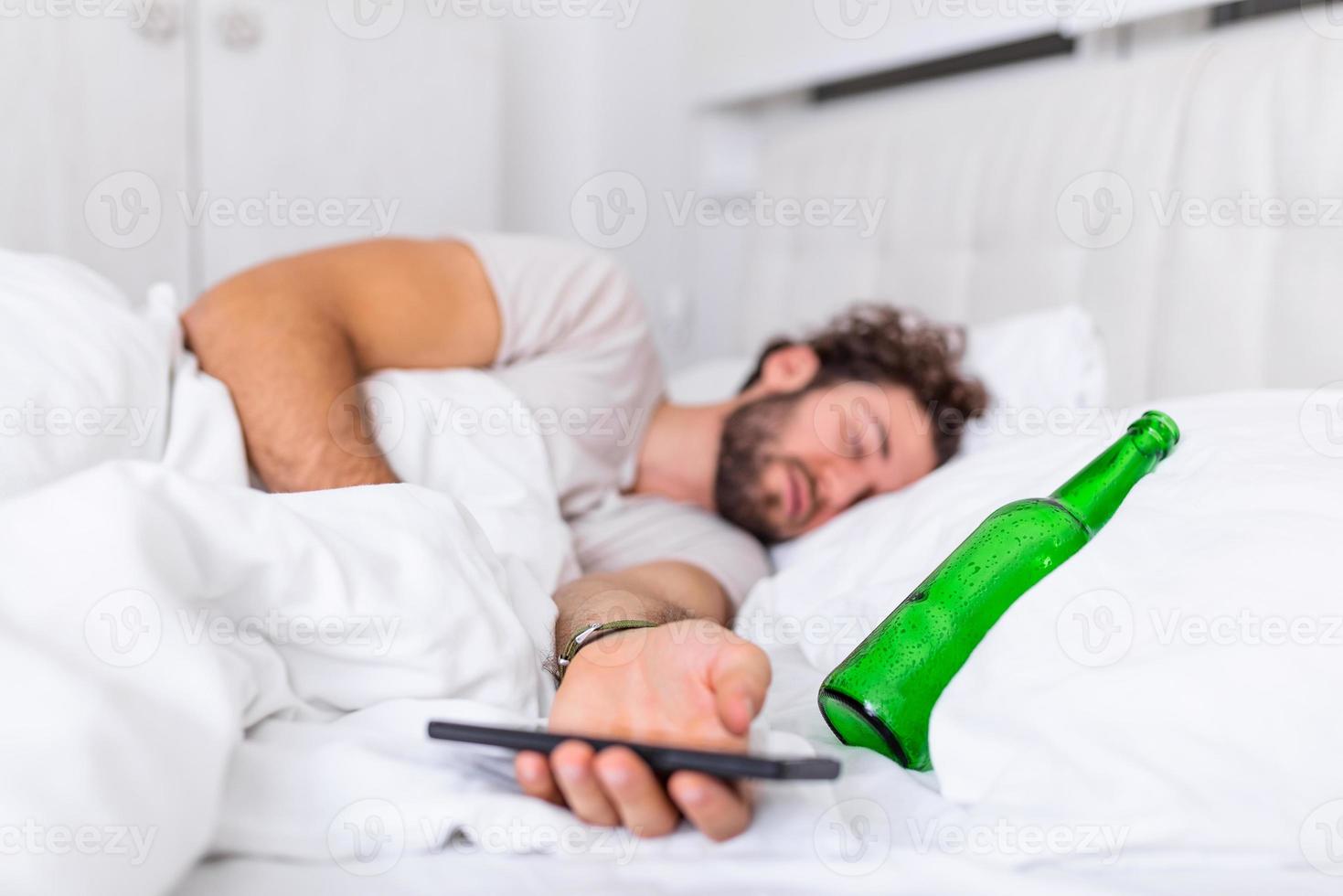 Texting while drunk concept. Young man lying in bed deadly drunken holding empty bottle of booze. intoxicated with alcohol . Alcoholism habitual drunkenness pernicious habit concept photo