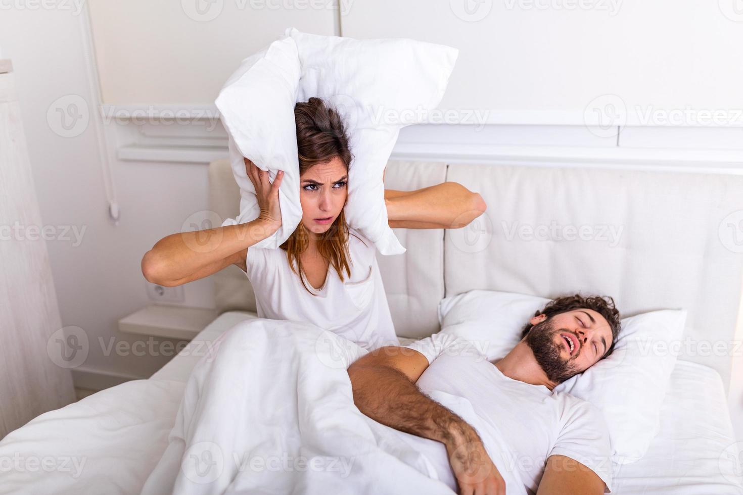 Man snoring while his wife is covering ears with pillow. Woman suffers from her male partner snoring in bed. Noise concept. Real people. Young irritated woman lying in bed with snoring husband photo