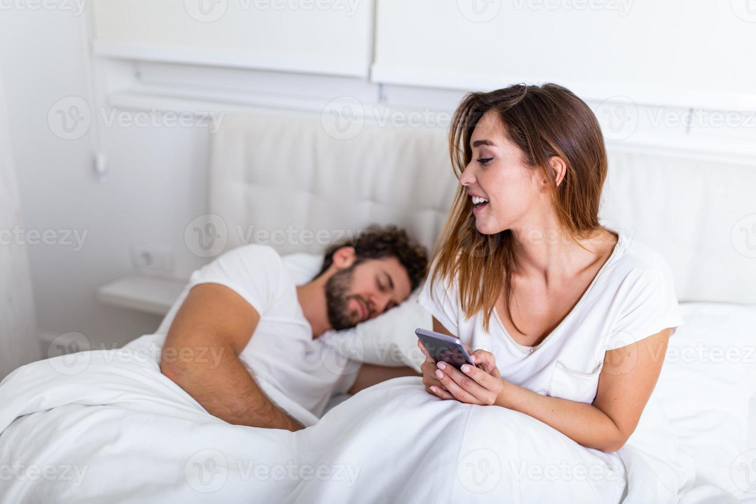 mujer mirando su teléfono mientras el hombre duerme en la cama. joven hermosa mujer hablando en privado por teléfono móvil mientras su marido duerme en la cama en el dormitorio foto