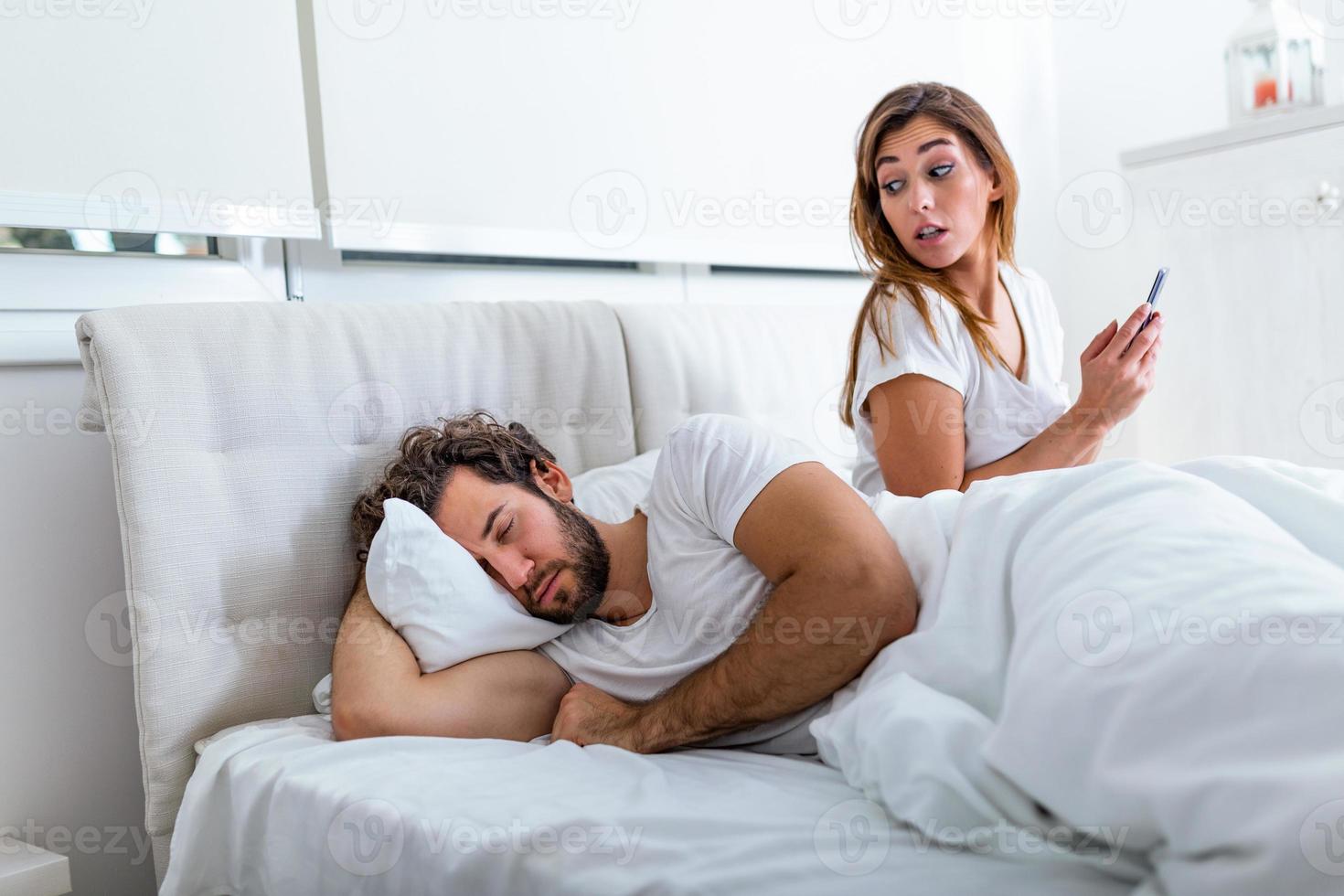Cheating wife using mobile phone lying in bed next to his sleeping husband. Affair. Cheating Girlfriend Chatting On Phone While Boyfriend Sleeping In Bedroom At Night. Selective Focus. photo