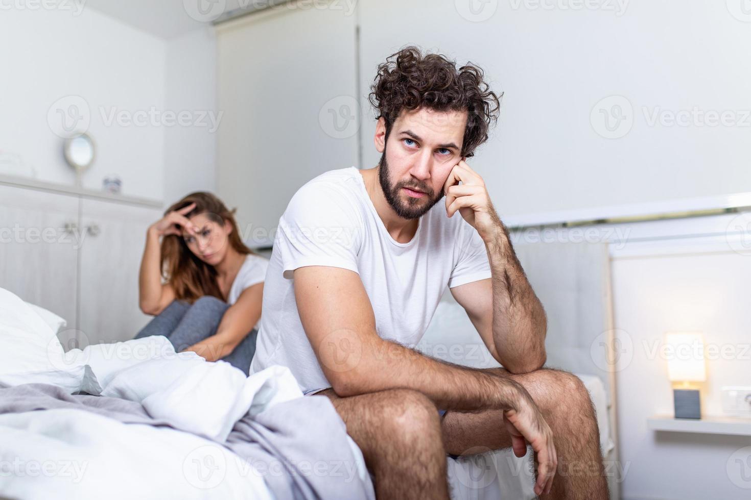 Young couple having a problem. Handsome man is leaning on his hand while sitting sadly on bed, woman is sitting in the background. Head full of thoughts when she is around photo