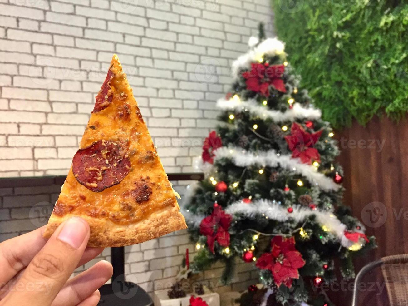 Hand held pizza slice with Christmas tree decoration background. photo