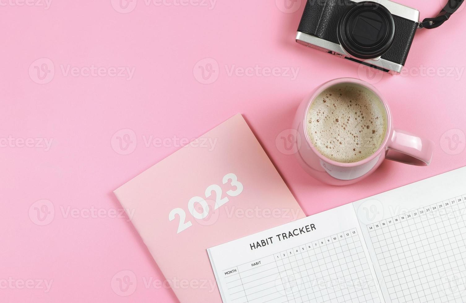 flat lay of habit tracker book on pink diary or planner 2023, pink cup of coffee and digital camera on pink background with copy space. photo