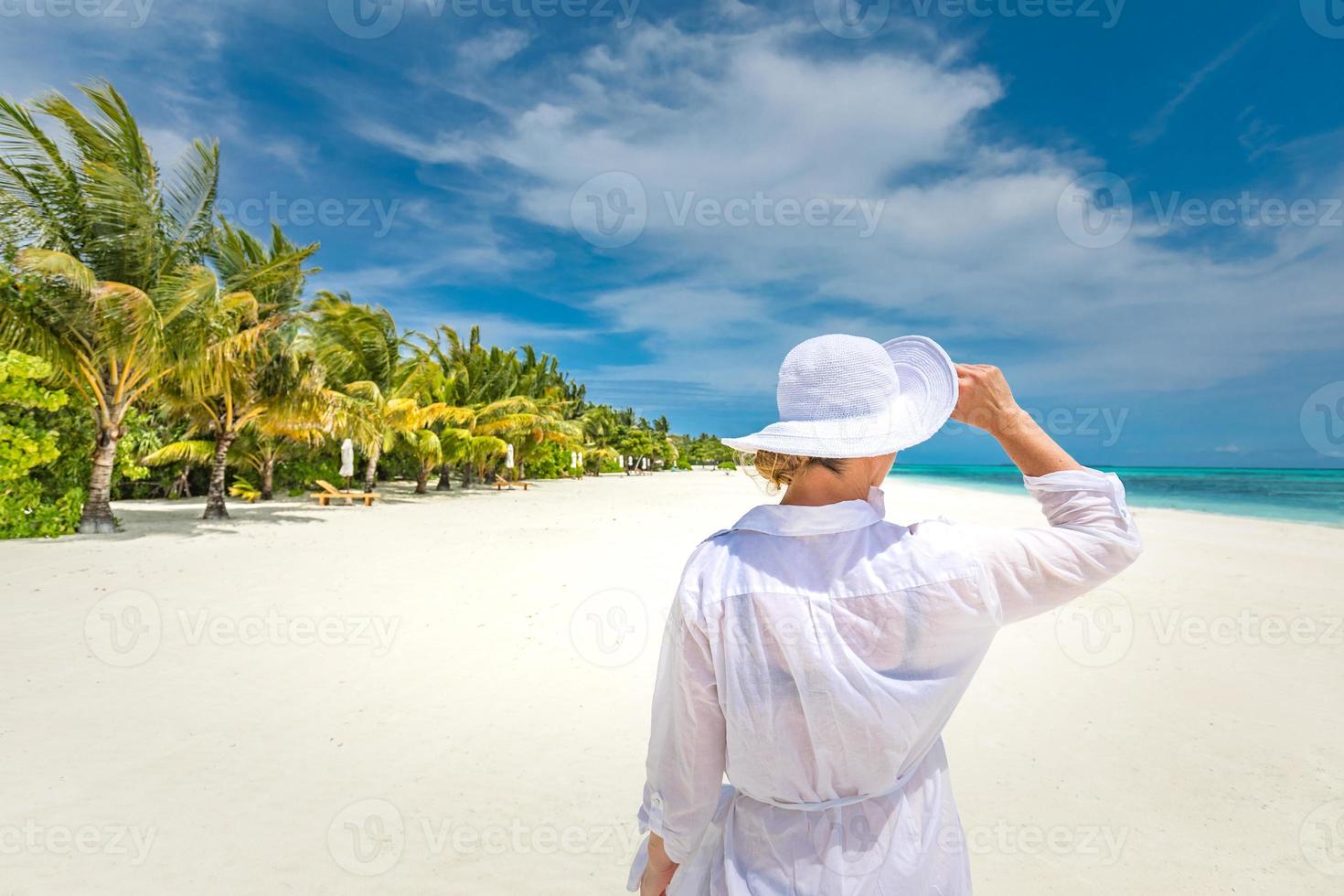 mujer viajera feliz con vestido blanco disfruta de sus vacaciones en la playa tropical y mira el mar. hermoso paisaje tropical, vacaciones de verano y concepto de turismo exótico. día soleado, humor feliz, relajación. foto