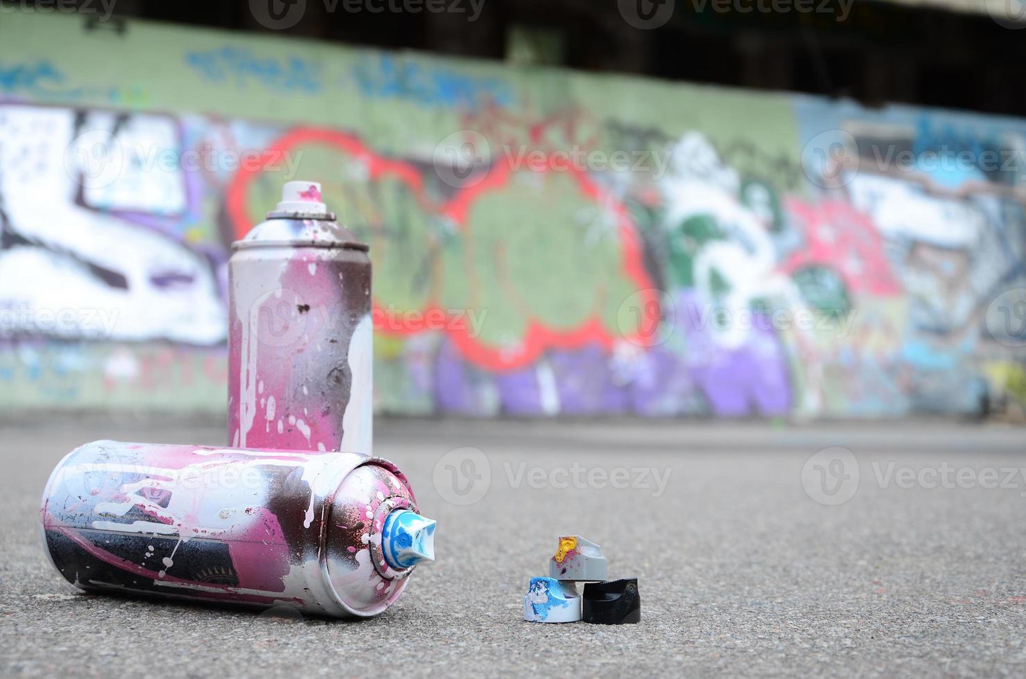 Several used spray cans with pink and white paint and caps for spraying paint under pressure is lies on the asphalt near the painted wall in colored graffiti drawings photo