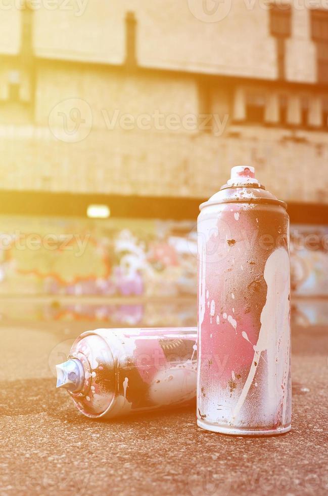 Several used spray cans with pink and white paint and caps for spraying paint under pressure is lies on the asphalt near the painted wall in colored graffiti drawings photo