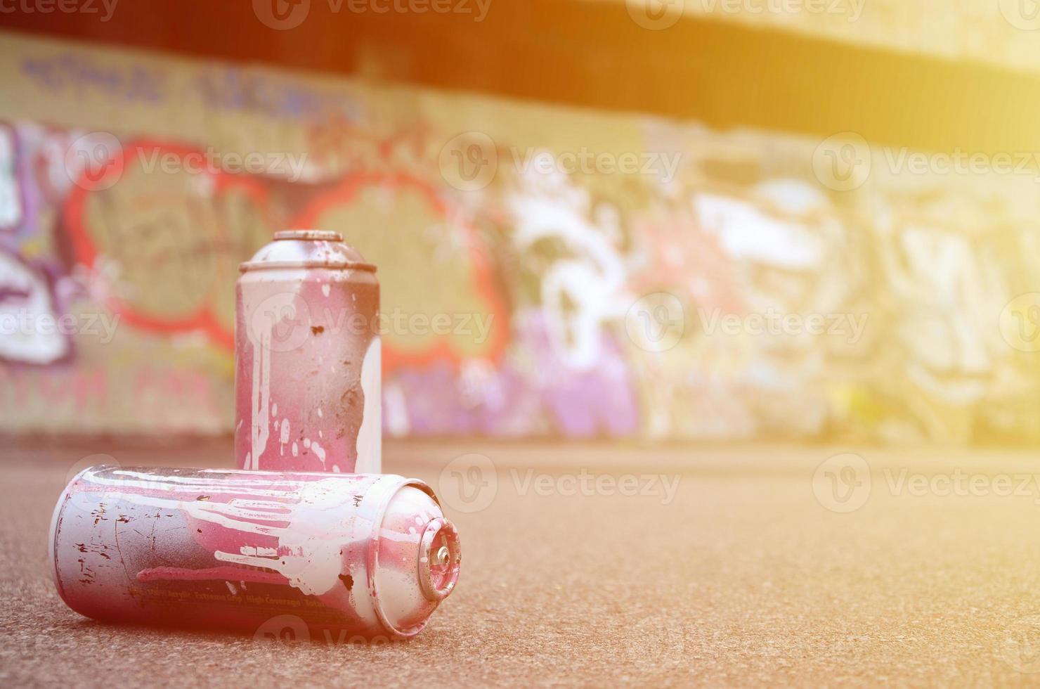 A few used spray cans with pink and white paint lie on the asphalt against the background of a painted wall in colorful graffiti drawings photo