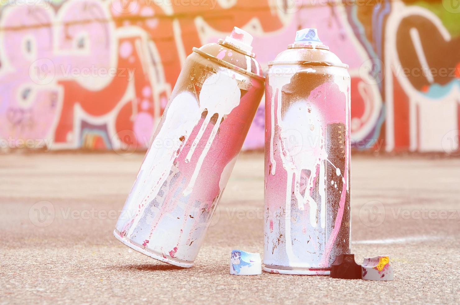 Several used spray cans with pink and white paint and caps for spraying paint under pressure is lies on the asphalt near the painted wall in colored graffiti drawings photo
