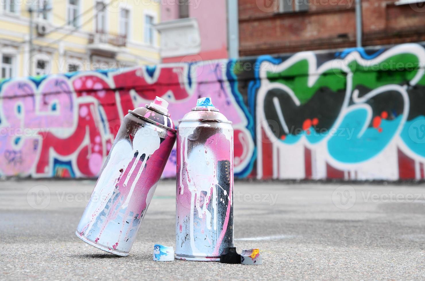 Several used spray cans with pink and white paint and caps for spraying paint under pressure is lies on the asphalt near the painted wall in colored graffiti drawings photo