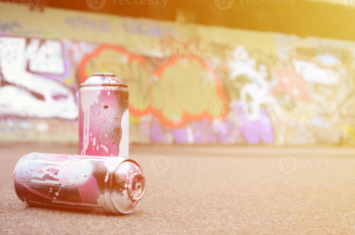 A few used spray cans with pink and white paint lie on the asphalt against the background of a painted wall in colorful graffiti drawings photo