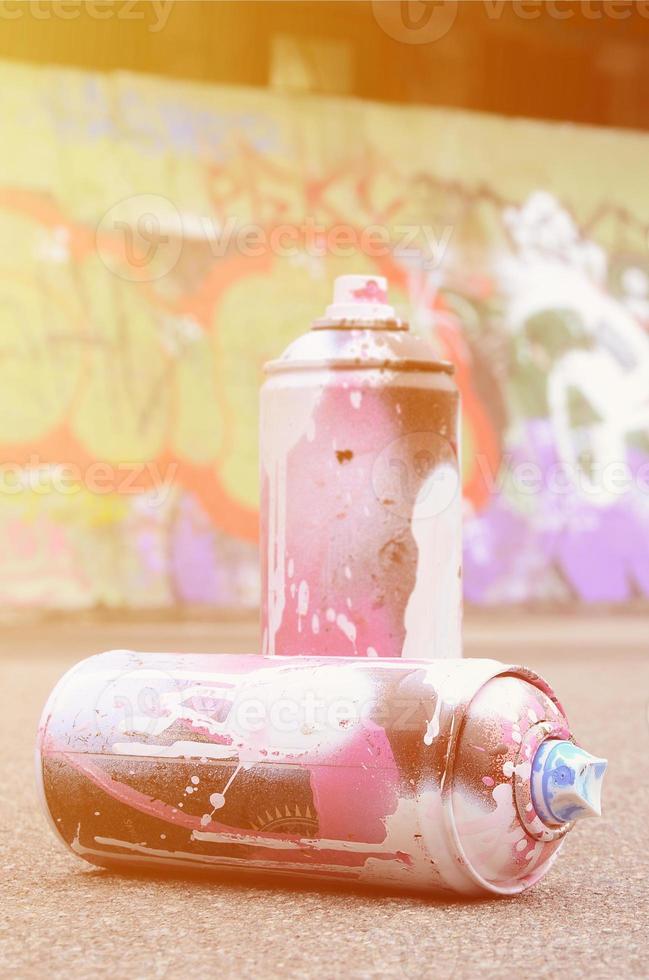 varias latas de aerosol usadas con pintura rosa y blanca y tapas para rociar pintura bajo presión se encuentran en el asfalto cerca de la pared pintada en dibujos de graffiti de colores foto