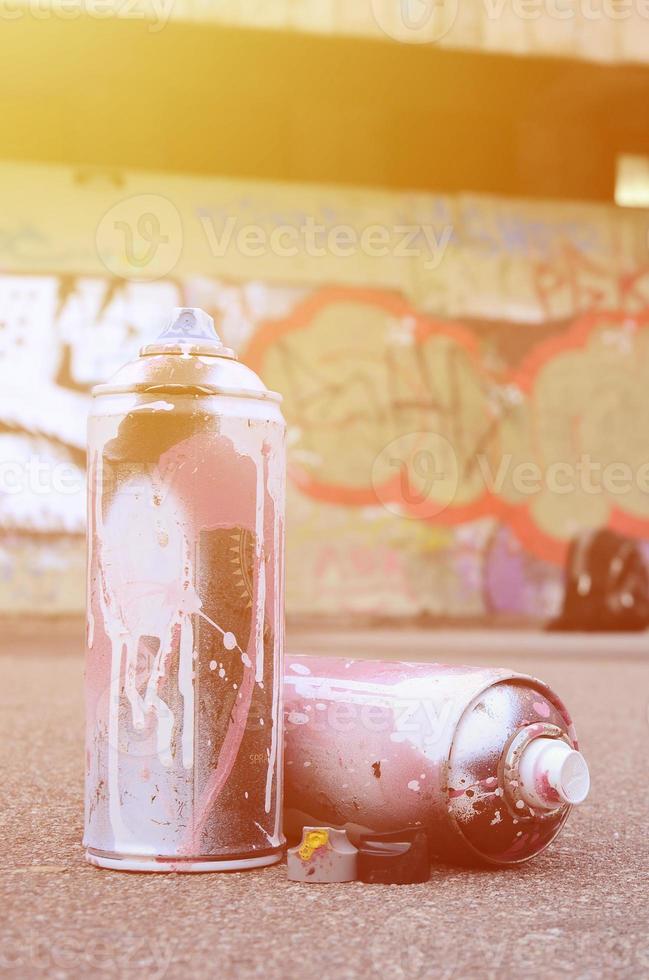 varias latas de aerosol usadas con pintura rosa y blanca y tapas para rociar pintura bajo presión se encuentran en el asfalto cerca de la pared pintada en dibujos de graffiti de colores foto