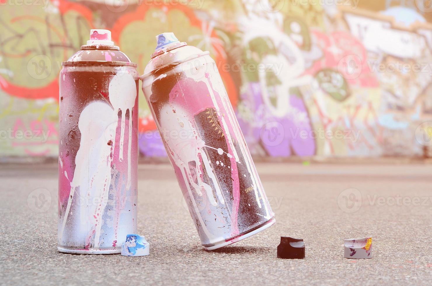 Several used spray cans with pink and white paint and caps for spraying paint under pressure is lies on the asphalt near the painted wall in colored graffiti drawings photo