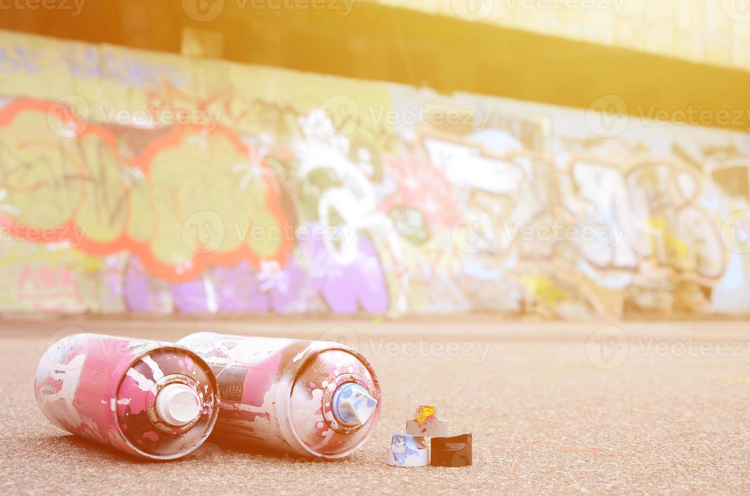 Several used spray cans with pink and white paint and caps for spraying paint under pressure is lies on the asphalt near the painted wall in colored graffiti drawings photo