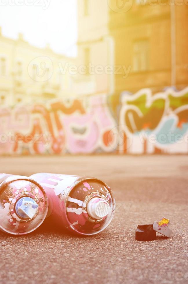 Several used spray cans with pink and white paint and caps for spraying paint under pressure is lies on the asphalt near the painted wall in colored graffiti drawings photo
