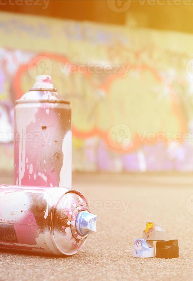 Several used spray cans with pink and white paint and caps for spraying paint under pressure is lies on the asphalt near the painted wall in colored graffiti drawings photo