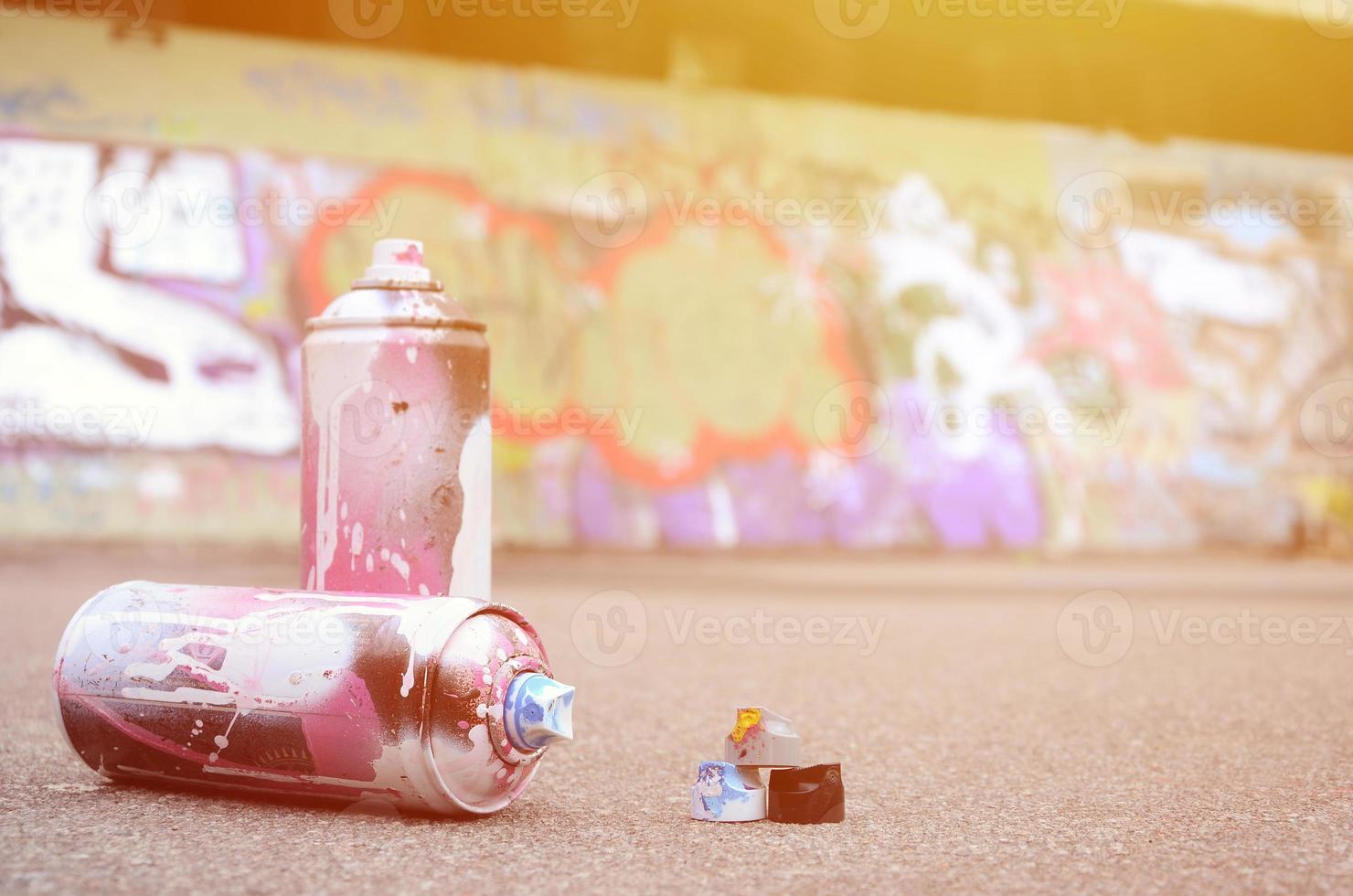 Several used spray cans with pink and white paint and caps for spraying paint under pressure is lies on the asphalt near the painted wall in colored graffiti drawings photo