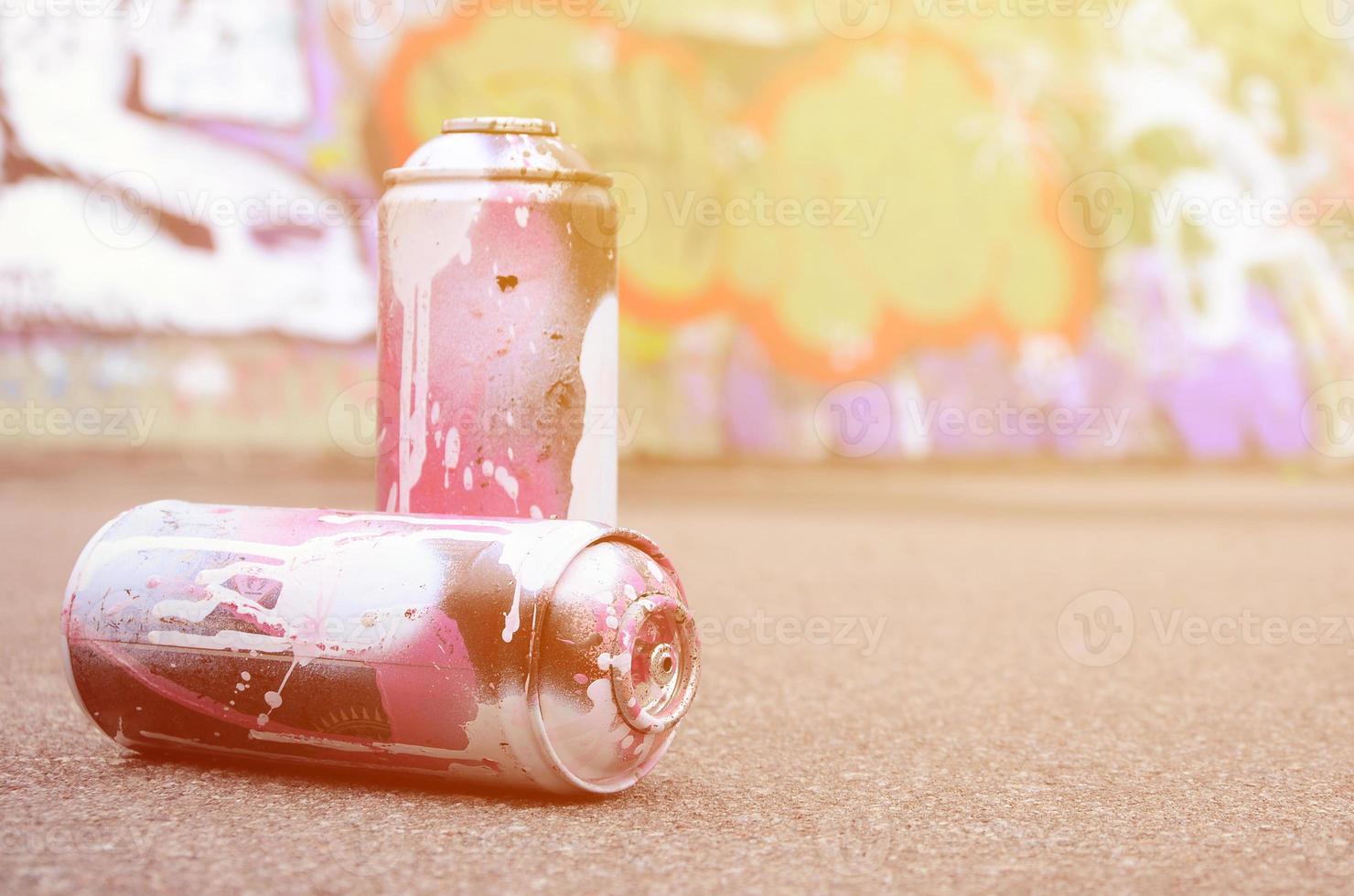 A few used spray cans with pink and white paint lie on the asphalt against the background of a painted wall in colorful graffiti drawings photo