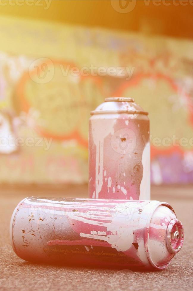 A few used spray cans with pink and white paint lie on the asphalt against the background of a painted wall in colorful graffiti drawings photo