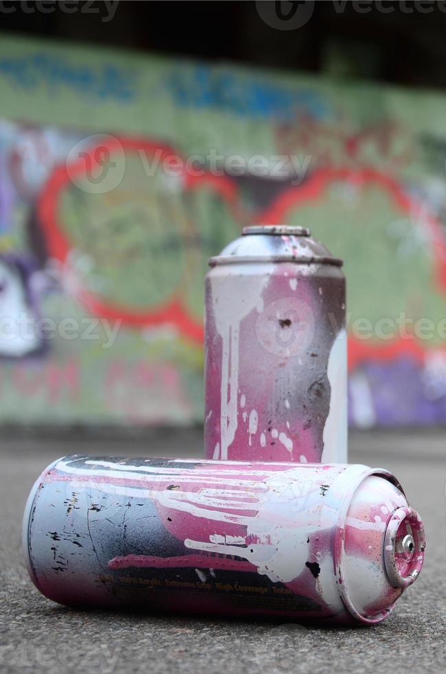 A few used spray cans with pink and white paint lie on the asphalt against the background of a painted wall in colorful graffiti drawings photo