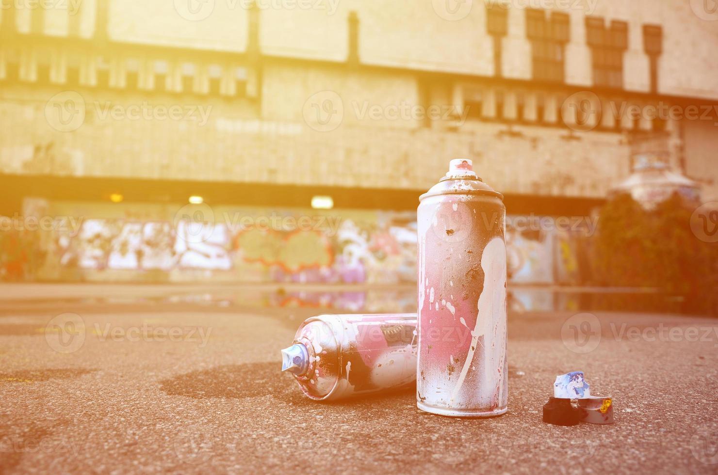 Several used spray cans with pink and white paint and caps for spraying paint under pressure is lies on the asphalt near the painted wall in colored graffiti drawings photo