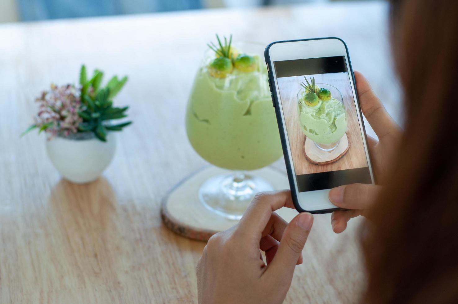 A woman in a coffee shop using a mobile phone to take pictures of avocado smoothie in front to upload photos to social media. Drink healthy day