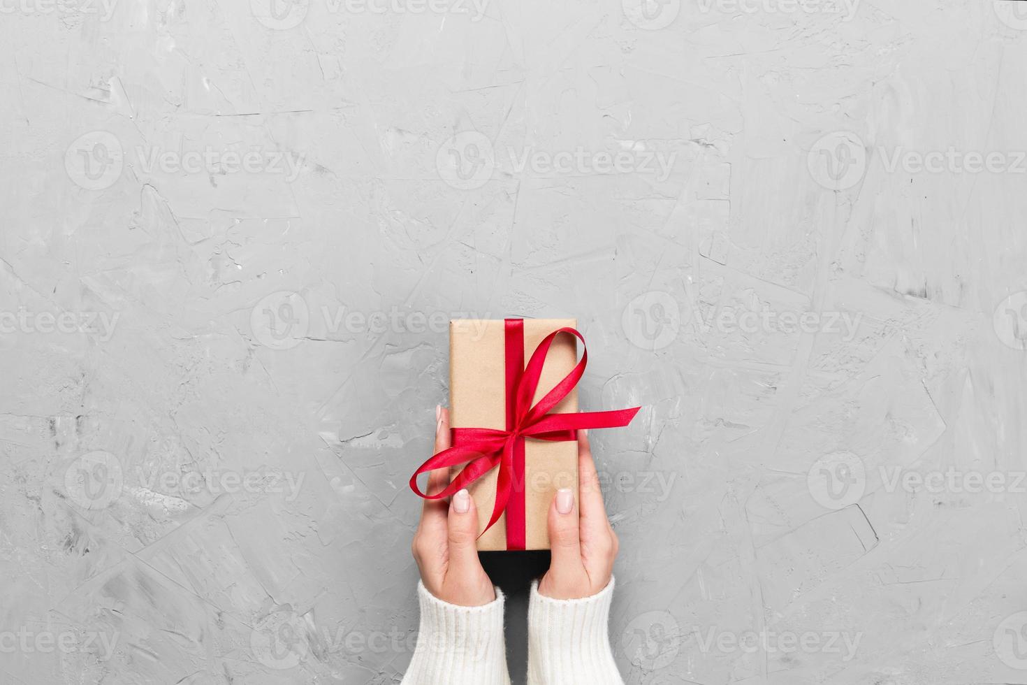 Woman hands give wrapped valentine or other holiday handmade present in paper with red ribbon. Present box, decoration of gift on white cement table, top view with copy space photo