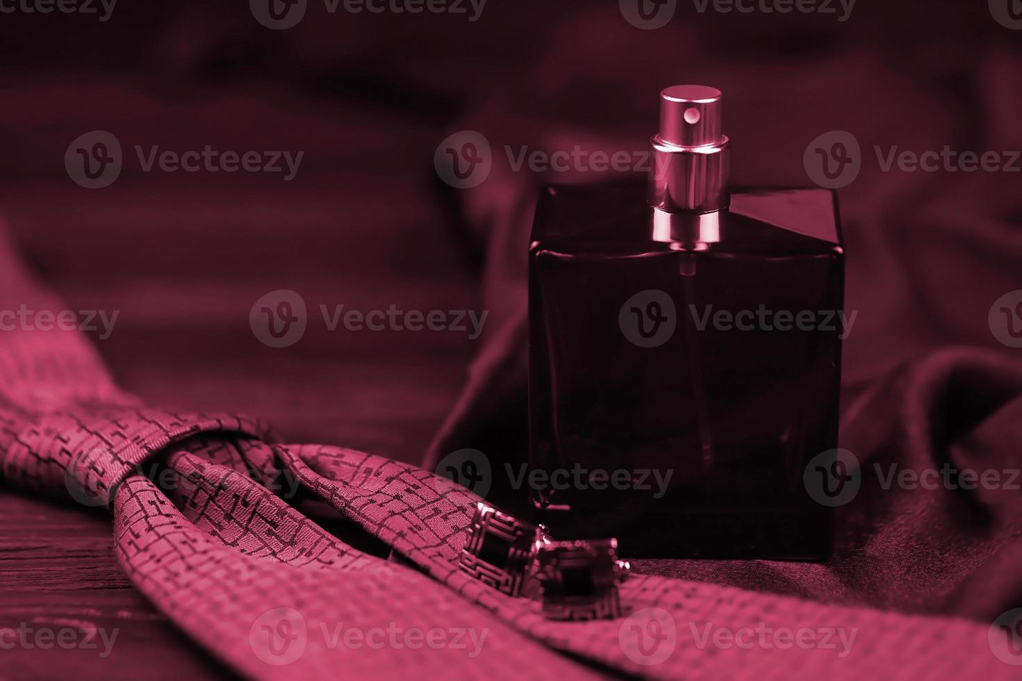 A bottle of mens cologne and cufflinks with blue tie lie on a black luxury fabric background on a wooden table. Mens accessories Image toned in Viva Magenta, color of the 2023 year photo