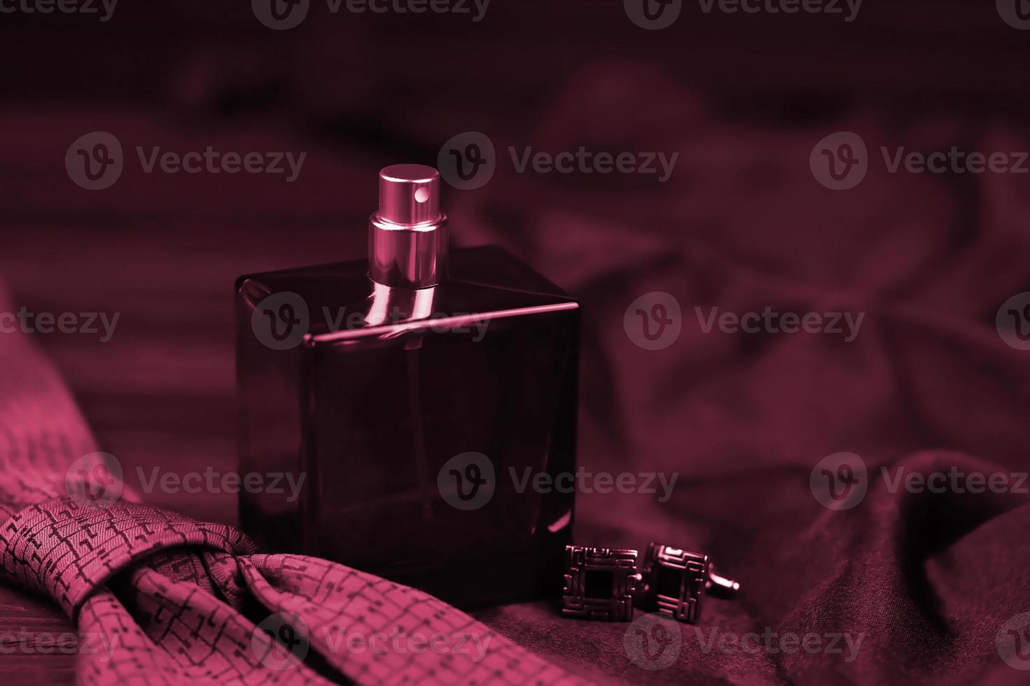 A bottle of mens cologne and cufflinks with blue tie lie on a black luxury fabric background on a wooden table. Mens accessories Image toned in Viva Magenta, color of the 2023 year photo