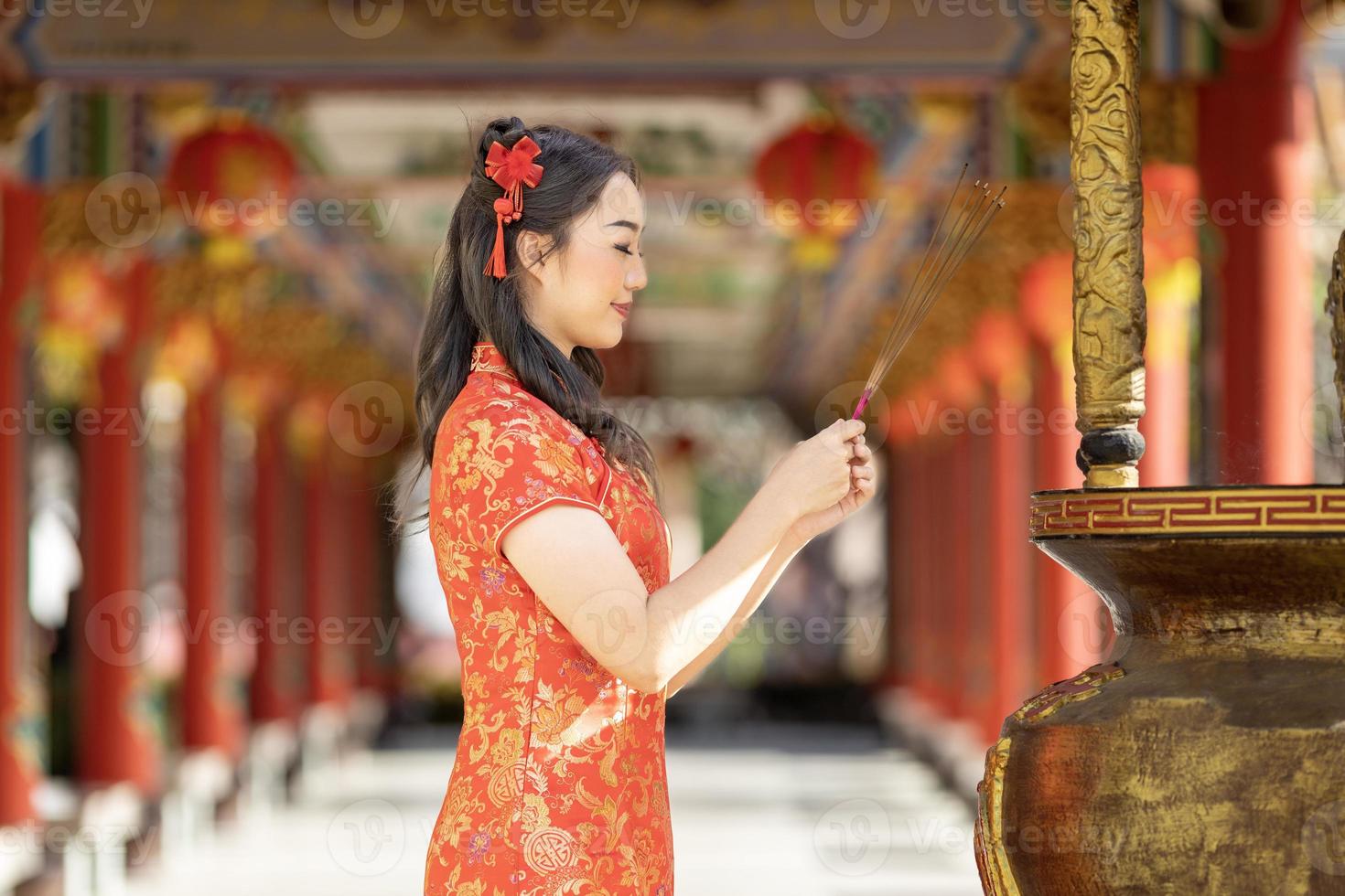 Asian woman in red cheongsam qipao dress is offering incense to the ancestral god inside Chinese Buddhist temple during lunar new year for  for best wish blessing and good luck concept photo