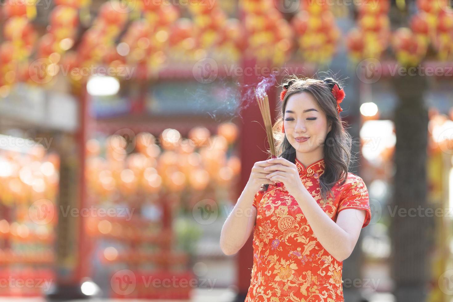 Asian woman in red cheongsam qipao dress is offering incense to the ancestral god inside Chinese Buddhist temple during lunar new year for  for best wish blessing and good luck concept photo