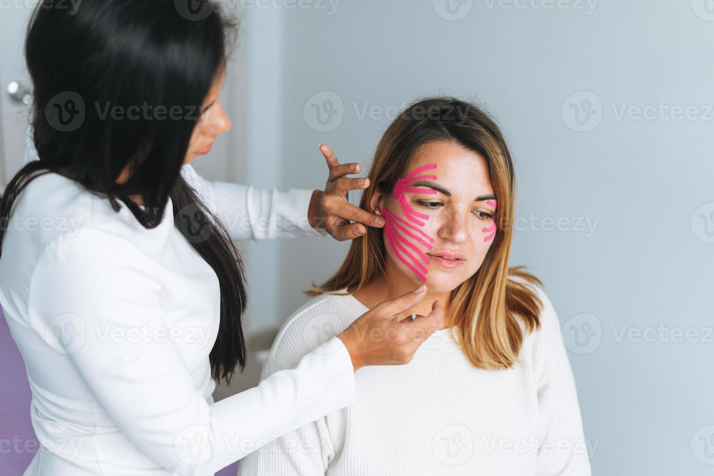 joven y hermosa mujer morena con el cosmetólogo médico de pelo largo hace cintas faciales para el paciente en el consultorio. las mujeres jóvenes realizan procedimientos antienvejecimiento con ayuda de cinta kinesio foto