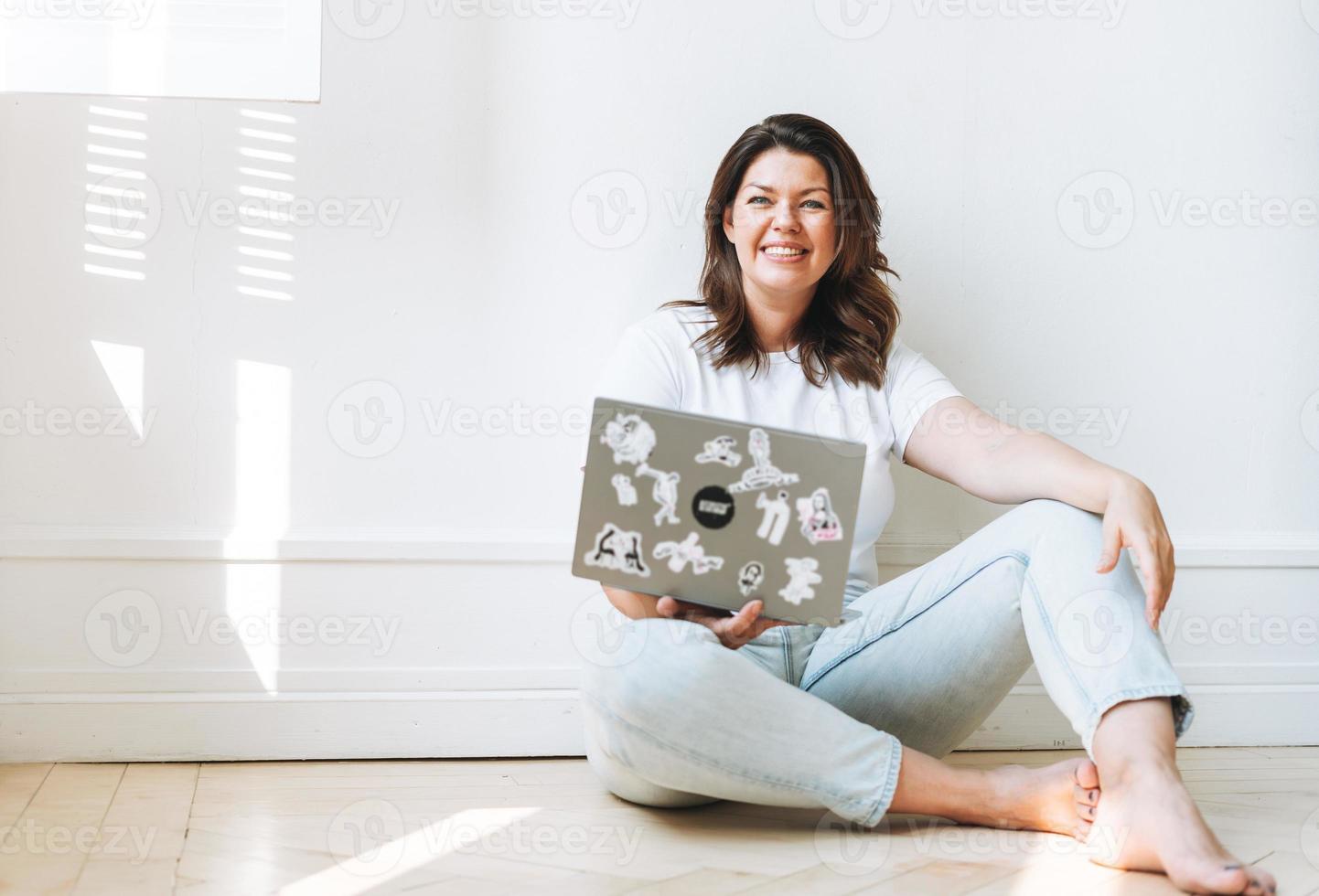 mujer morena sonriente con ropa informal y cuerpo positivo de talla grande usando una laptop sentada en el suelo en una habitación luminosa foto