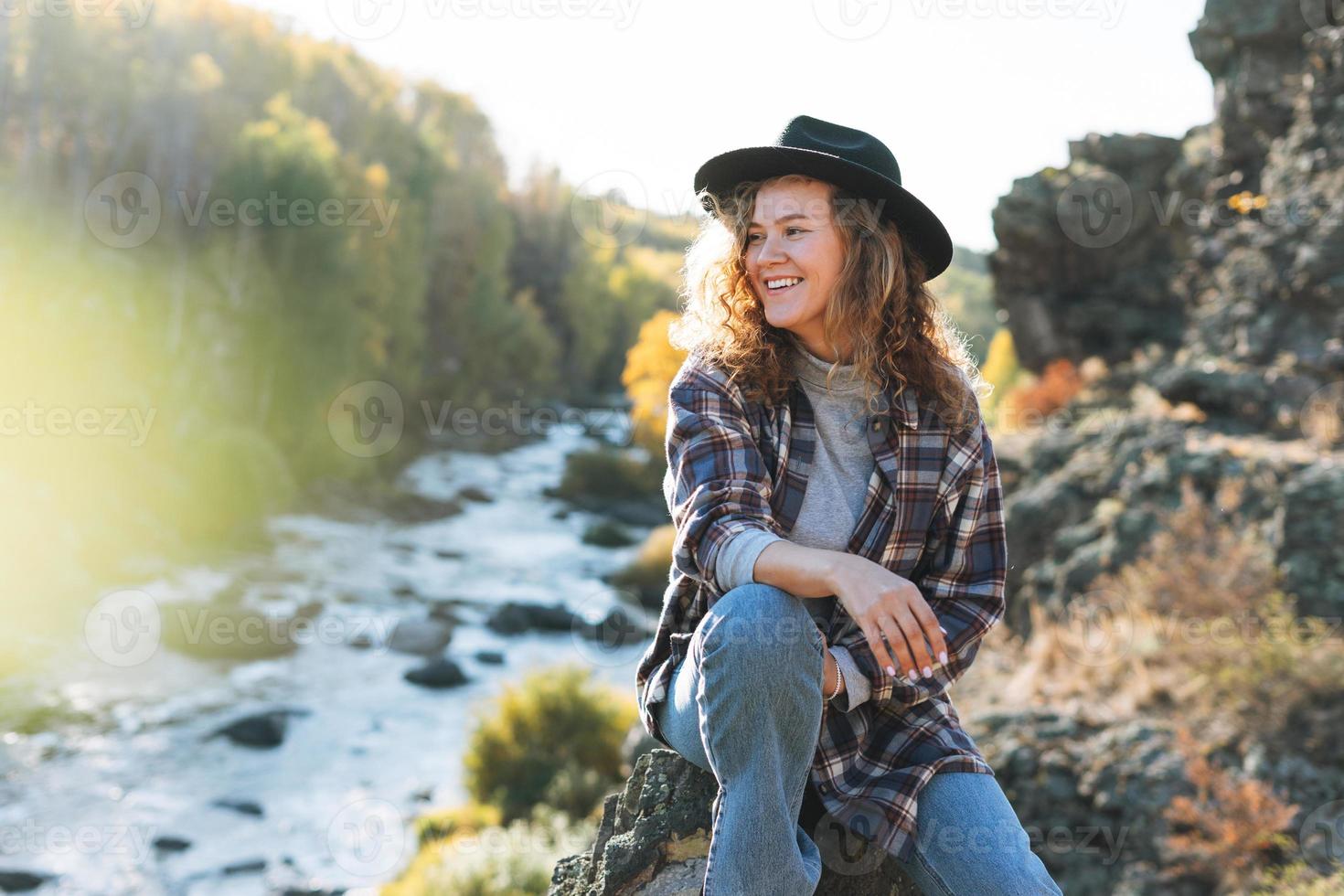 joven mujer hermosa con cabello rizado en sombrero de fieltro y camisa a cuadros en jeans mira la vista mágica de las montañas y el río, caminatas en la naturaleza otoñal, ambiente sostenible foto