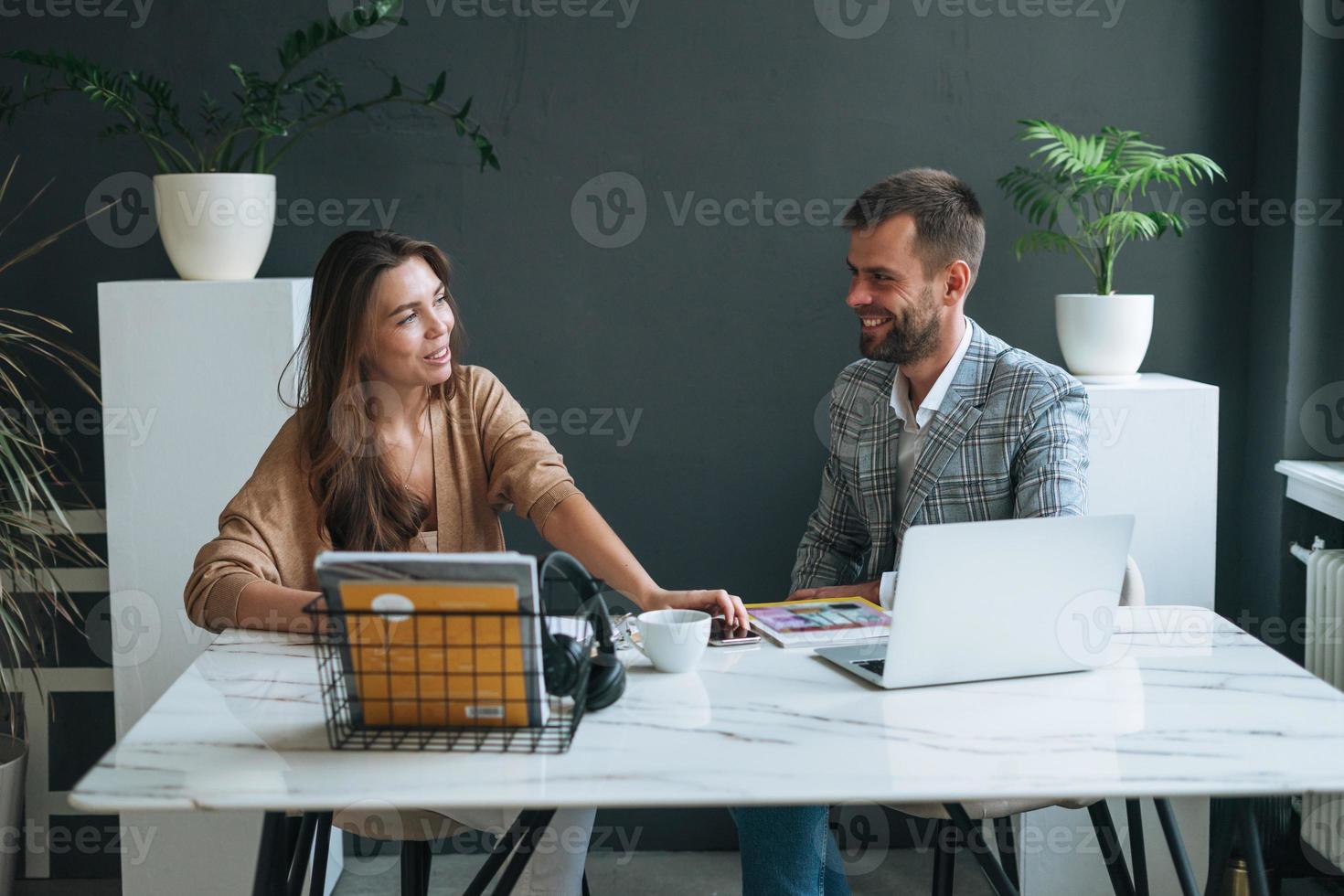 Young brunette woman with long hair in stylish suit working with young man using laptop on table with green house plant in bright modern office, team work photo