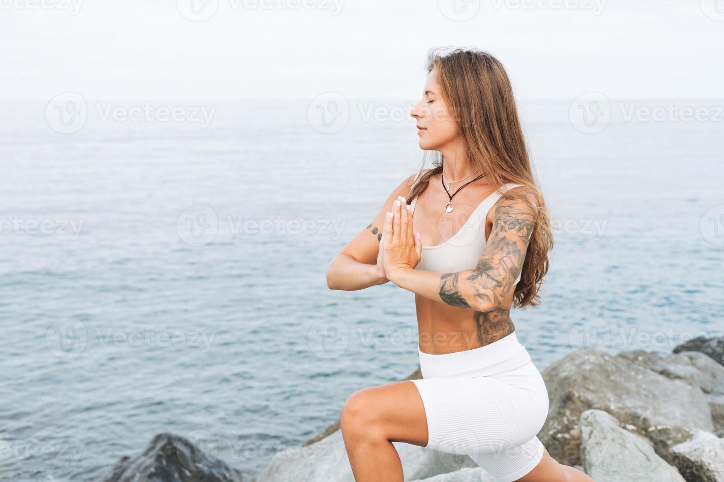 Una mujer joven y hermosa con el pelo largo y ropa deportiva blanca practica yoga y meditación en la playa del mar foto