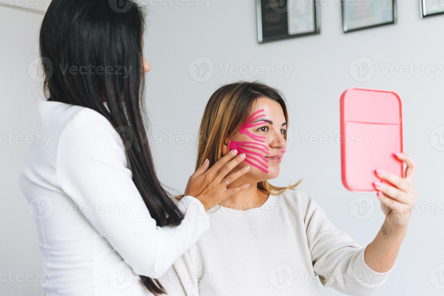 joven y hermosa mujer morena con el médico cosmetólogo de pelo largo hace cintas faciales para el paciente en el consultorio. las mujeres jóvenes realizan procedimientos antienvejecimiento con la ayuda de kinesio tape foto