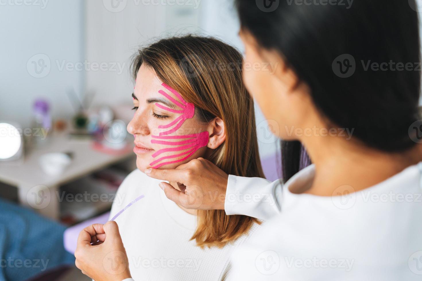 joven y hermosa mujer morena con el cosmetólogo médico de pelo largo hace cintas faciales para el paciente en el consultorio. las mujeres jóvenes realizan procedimientos antienvejecimiento con ayuda de cinta kinesio foto